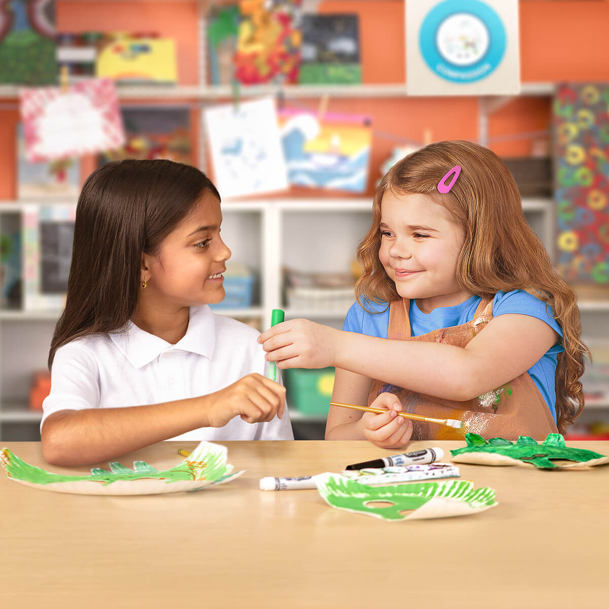 Two young girls smile and share art supplies while painting green paper crafts together at a classroom table.