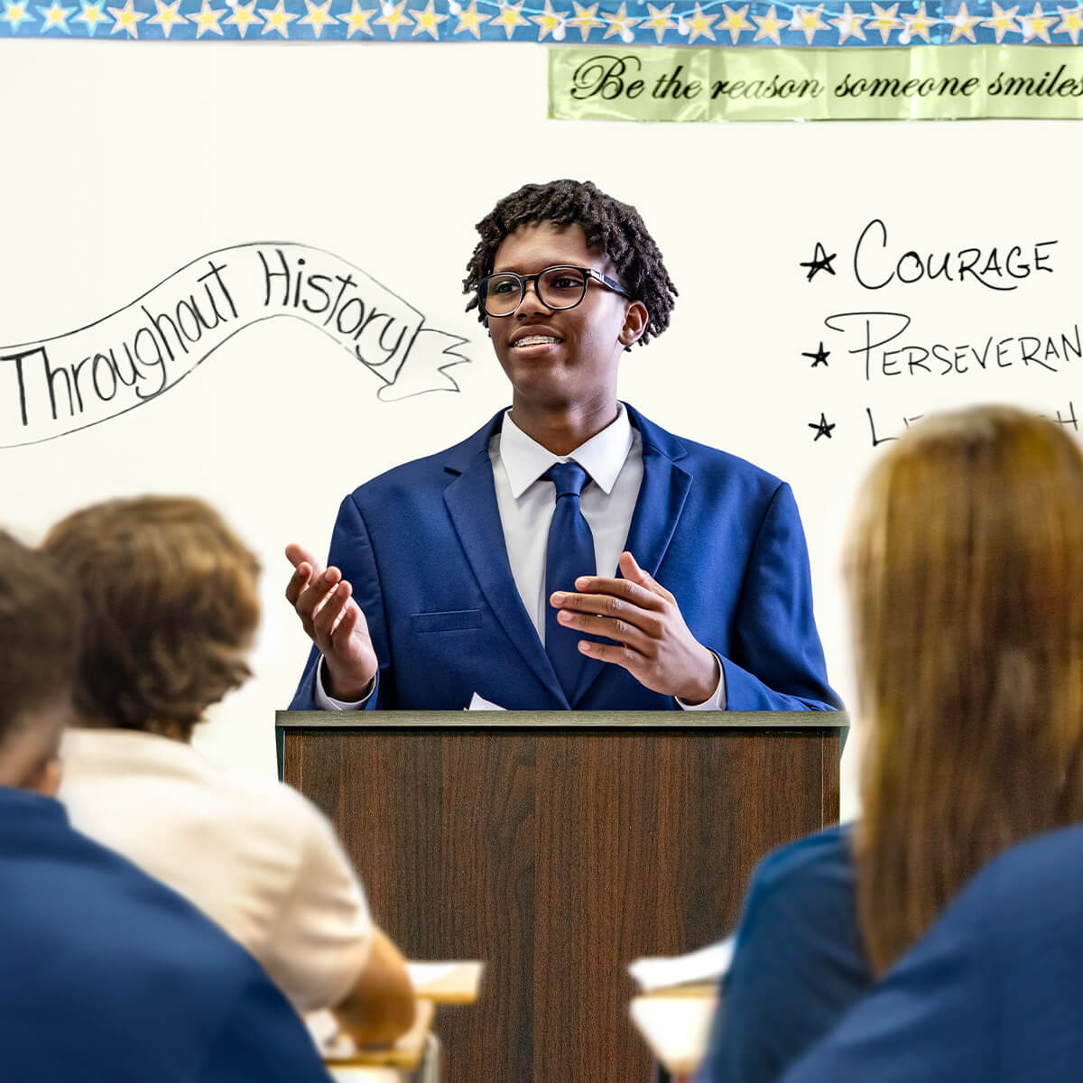 Student presenter gestures while speaking to peers, framed by classroom messages about courage and perseverance.