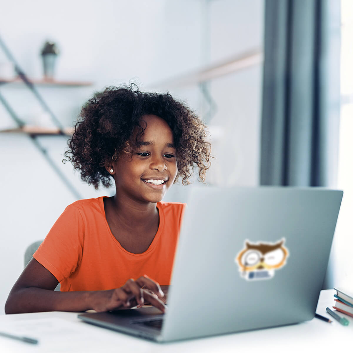 Young student works happily on a laptop in a bright home setting, wearing an orange T-shirt at a tidy desk.